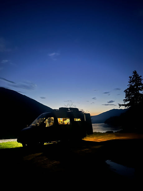 Cozy RV rental parked by a mountain lake at dusk. Warm lights glow inside the campervan.