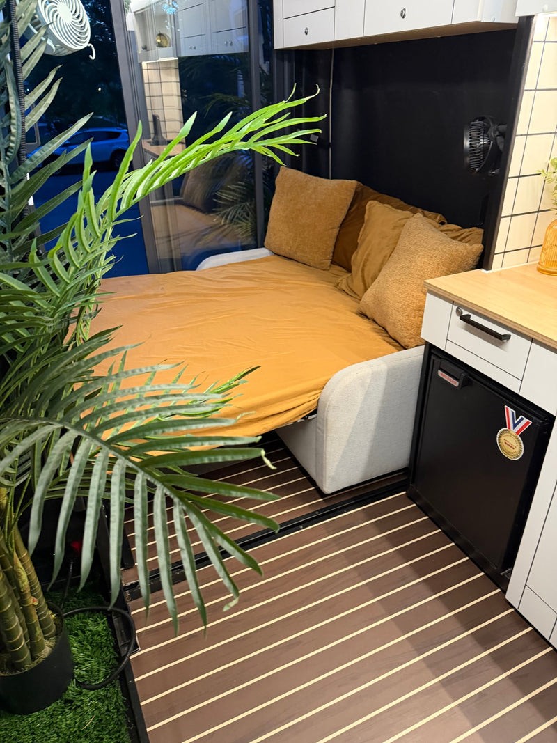 This rv rental features a compact living space with a pull-out bed, golden sheets, cushions, mini-fridge, and faux palm plant. — photo 9