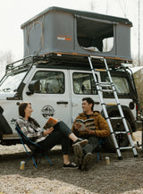 Two people relax by their RV rental, a Jeep Wrangler with a Roofnest rooftop tent, perfect for an Alaska camping adventure.
