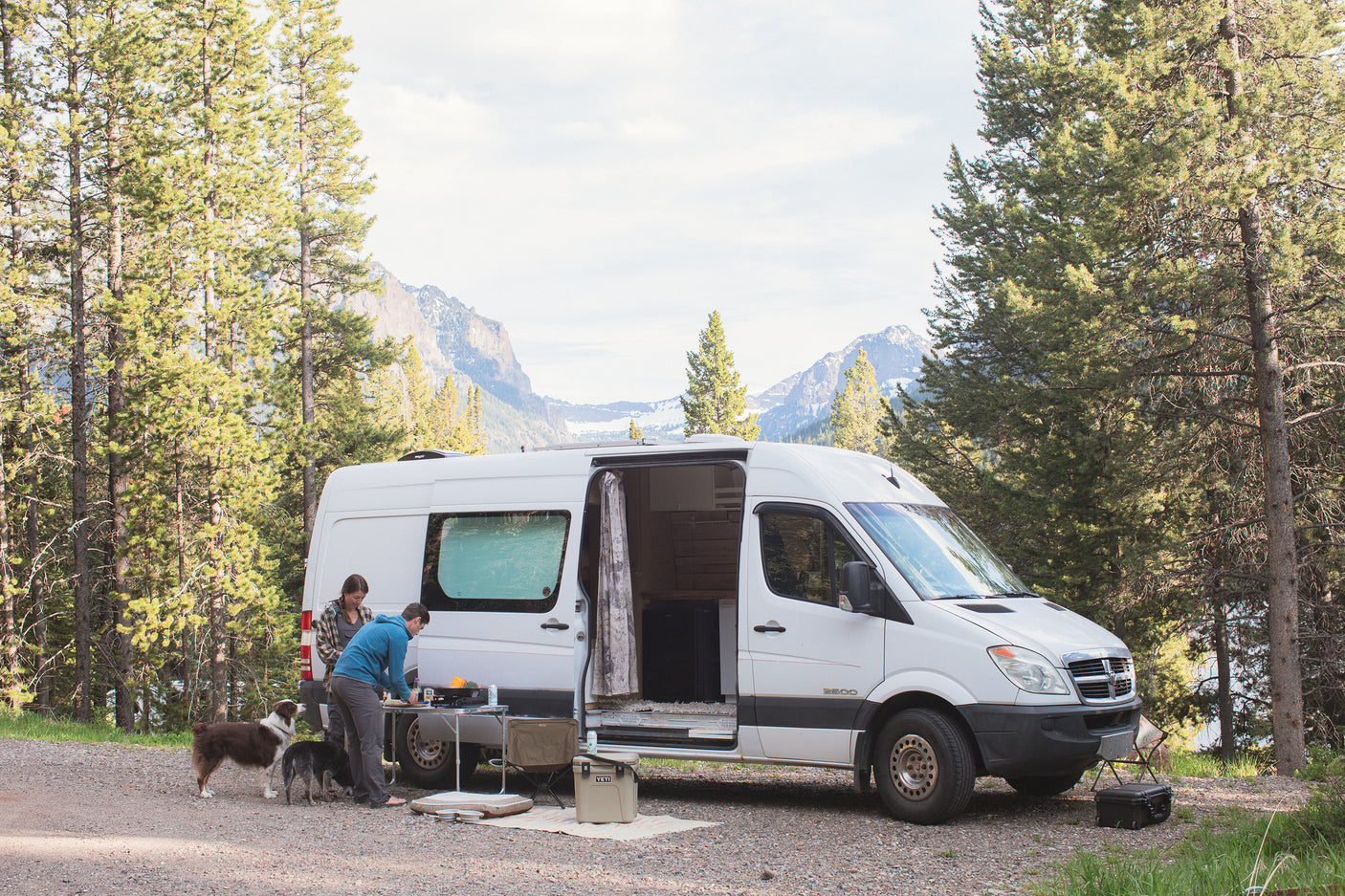 RV rental adventure: Two people and their dogs prepare food outside a white camper van in a mountain forest setting.
