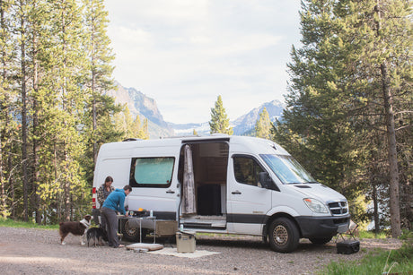 RV rental adventure: Two people and their dogs prepare food outside a white camper van in a mountain forest setting.