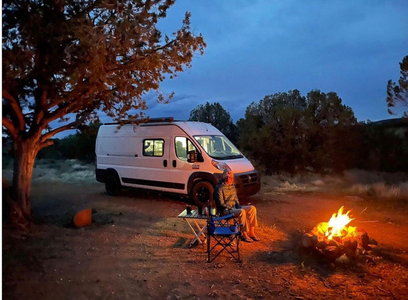 RV rental van parked at a rustic dusk camp. Person by glowing campfire illuminates the foreground near a tree.