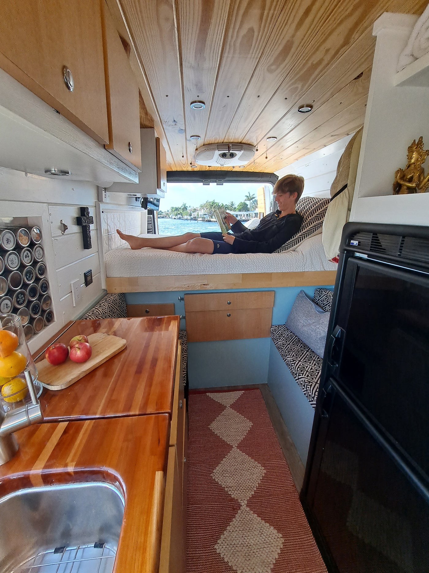 A cozy RV rental interior: A person sits on a bed by a window with a water view. Wood paneling, kitchen, and a Buddha statue complete the scene. — photo 15
