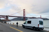 White camper van (RV rental) by San Francisco's iconic Golden Gate Bridge, with people strolling along the waterfront.