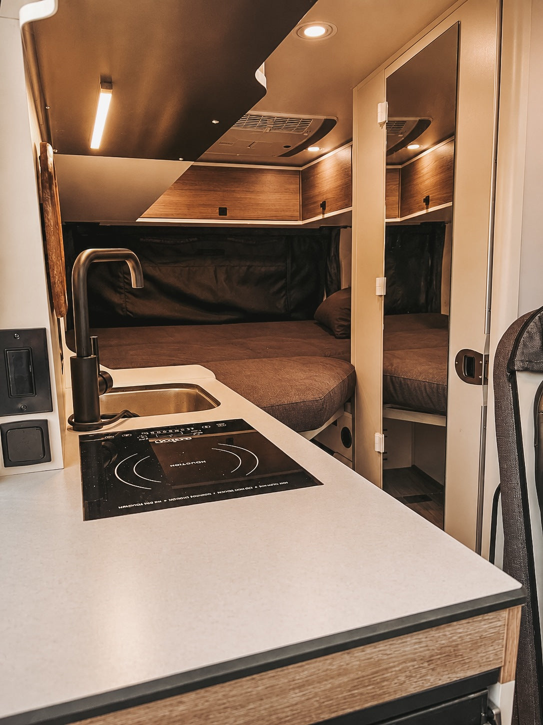 RV rental interior: A modern kitchenette with sink & induction cooktop is left, facing a cozy bed with dark bedding and mirrored storage.
