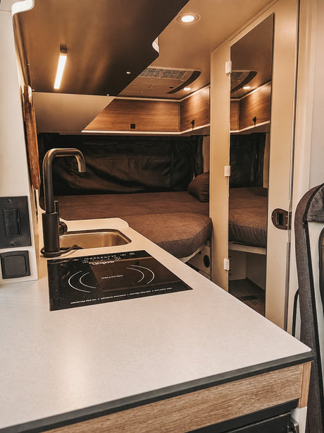 RV rental interior: A modern kitchenette with sink & induction cooktop is left, facing a cozy bed with dark bedding and mirrored storage.