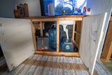 Compact RV rental interior reveals under-sink water system: fresh water input, grey water collection, and storage tanks.