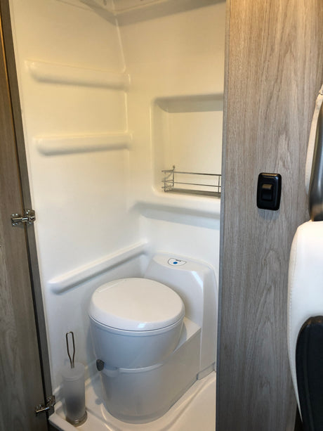 Inside an RV rental: a compact bathroom featuring a white toilet, molded shower walls with shelves, and a metal caddy.