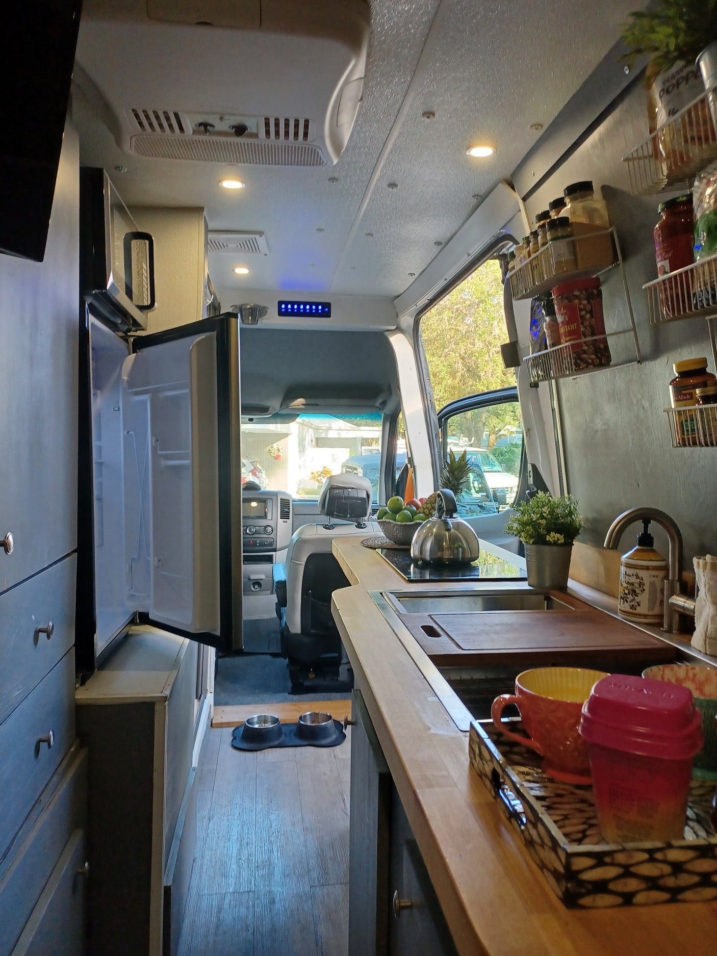 View inside an RV rental camper van: kitchenette with open fridge, sink, organized spices, and fruit. Compact and functional travel space.