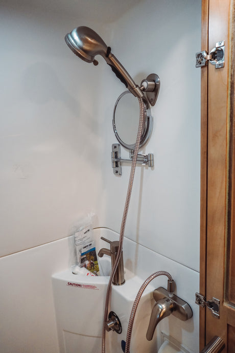 Inside a compact RV rental bathroom, showing a shower, sink, adjustable mirror, and various controls.