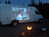 Cozy rv rental movie night: A film projected onto the white van, featuring a man on screen, with a warm campfire.