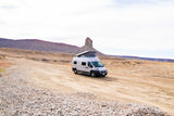 White pop-top RV rental on a dirt road, amidst stunning desert mountains and unique rock formations. Ready for adventure.