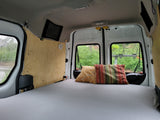 RV rental interior: A comfy bed with colorful pillows in a camper van, overlooking lush greenery through the back windows.
