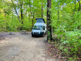 RV rental pop-top camper van at a shaded forest campsite with a fire pit and picnic table, surrounded by lush green trees.