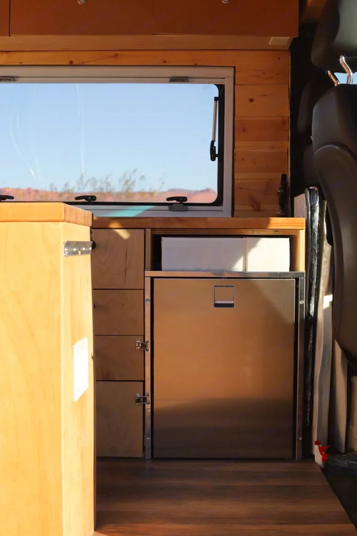 RV rental kitchenette with built-in stainless steel fridge, wood cabinets, and desert window view. Van life comfort.