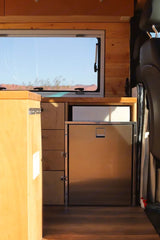 RV rental kitchenette with built-in stainless steel fridge, wood cabinets, and desert window view. Van life comfort.