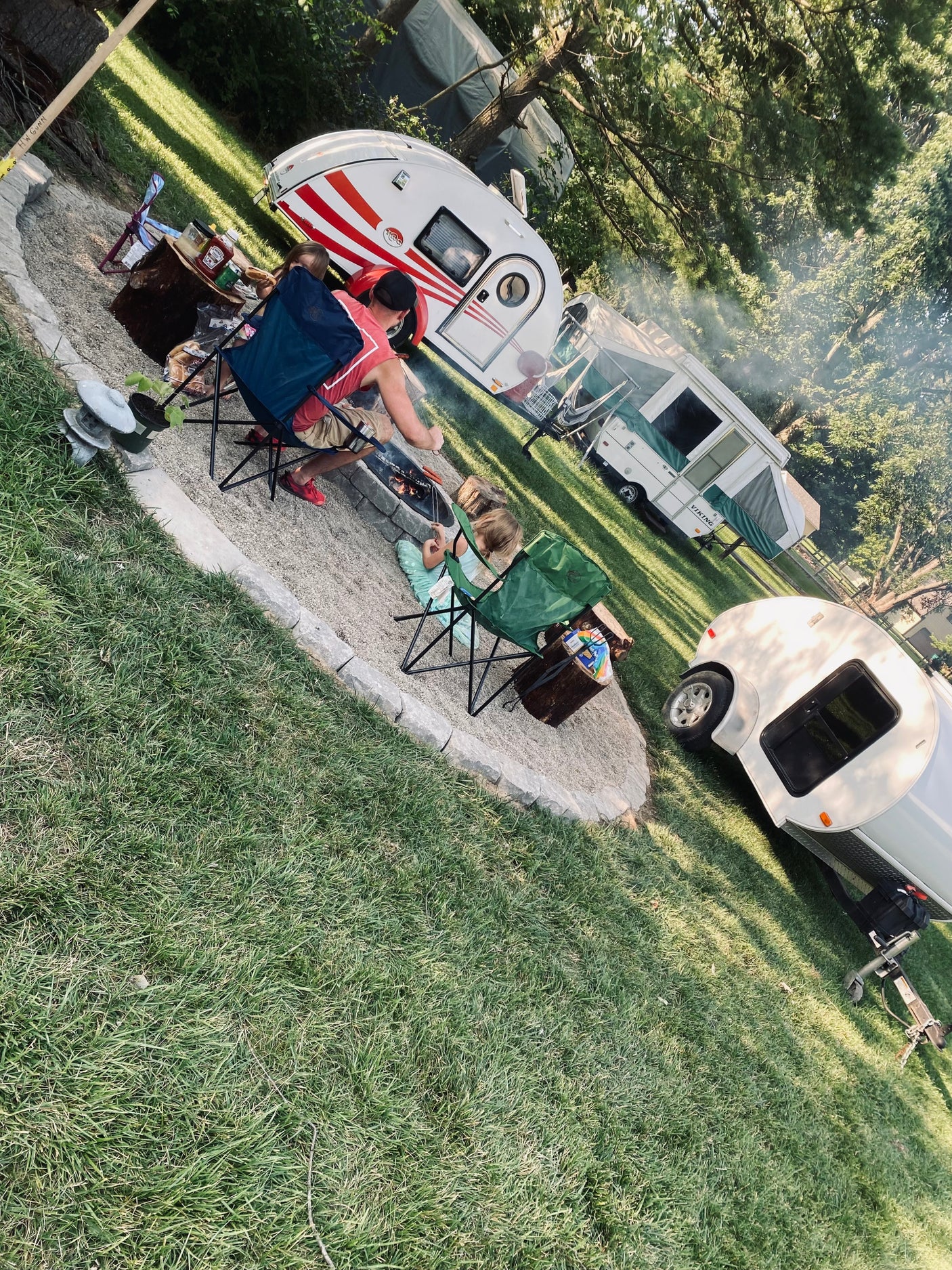 Campfire cookout with a family enjoying their RV rental setup: a white teardrop trailer and a green pop-up camper.