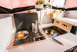 A compact rv rental kitchen is ready for breakfast. A pancake cooks on the Dometic stove, next to a coffee pot and sink.