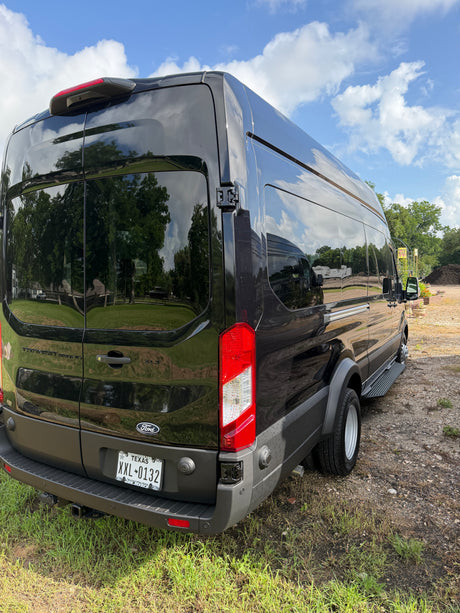 Black Ford Transit van for RV rental, detailed from the rear, reflecting sky & trees on its sleek surface.