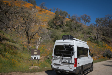White RV rental van parked on a dirt road by a Sierra National Forest sign, surrounded by hillsides vibrant with orange wildflowers.