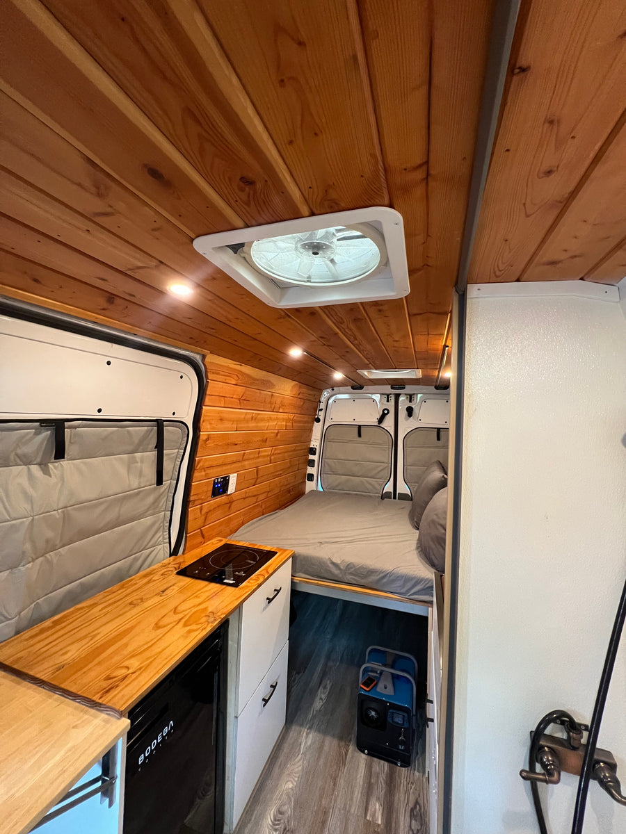 Camper van RV rental interior: wooden ceiling, cozy bed, kitchen with induction cooktop, fridge, and portable power station.