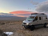 An RV rental, a grey Mercedes-Benz camper van, parked in a vast desert landscape with mountains under a vibrant sunset.