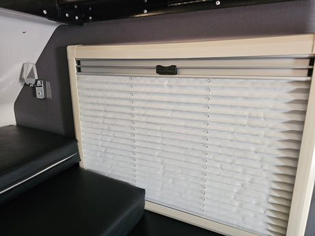 RV rental interior: closed light gray pleated blinds on a window, beige frame. Black cushions below in a compact camper area.