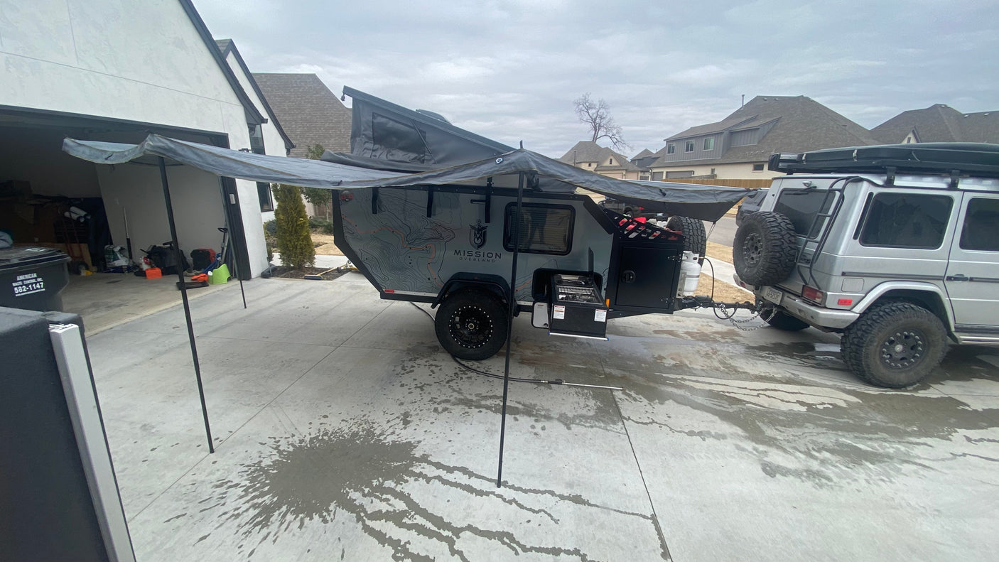 This **RV rental** setup features an overland trailer with an extended awning and outdoor kitchen, hitched to a G-Wagon in a driveway.