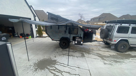 This **RV rental** setup features an overland trailer with an extended awning and outdoor kitchen, hitched to a G-Wagon in a driveway.
