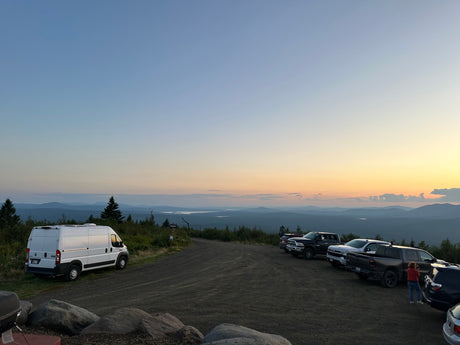 Ram ProMaster RV rental parked at a mountain overlook with a sunset sky over distant lakes and mountains.|A white RV rental camper van is parked on a scenic mountain road under a bright blue sky