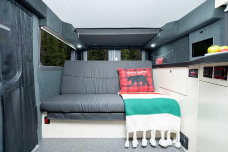 Experience comfort in this RV rental! Interior features a comfy bench, plaid pillow, blanket, and overhead bed/storage.