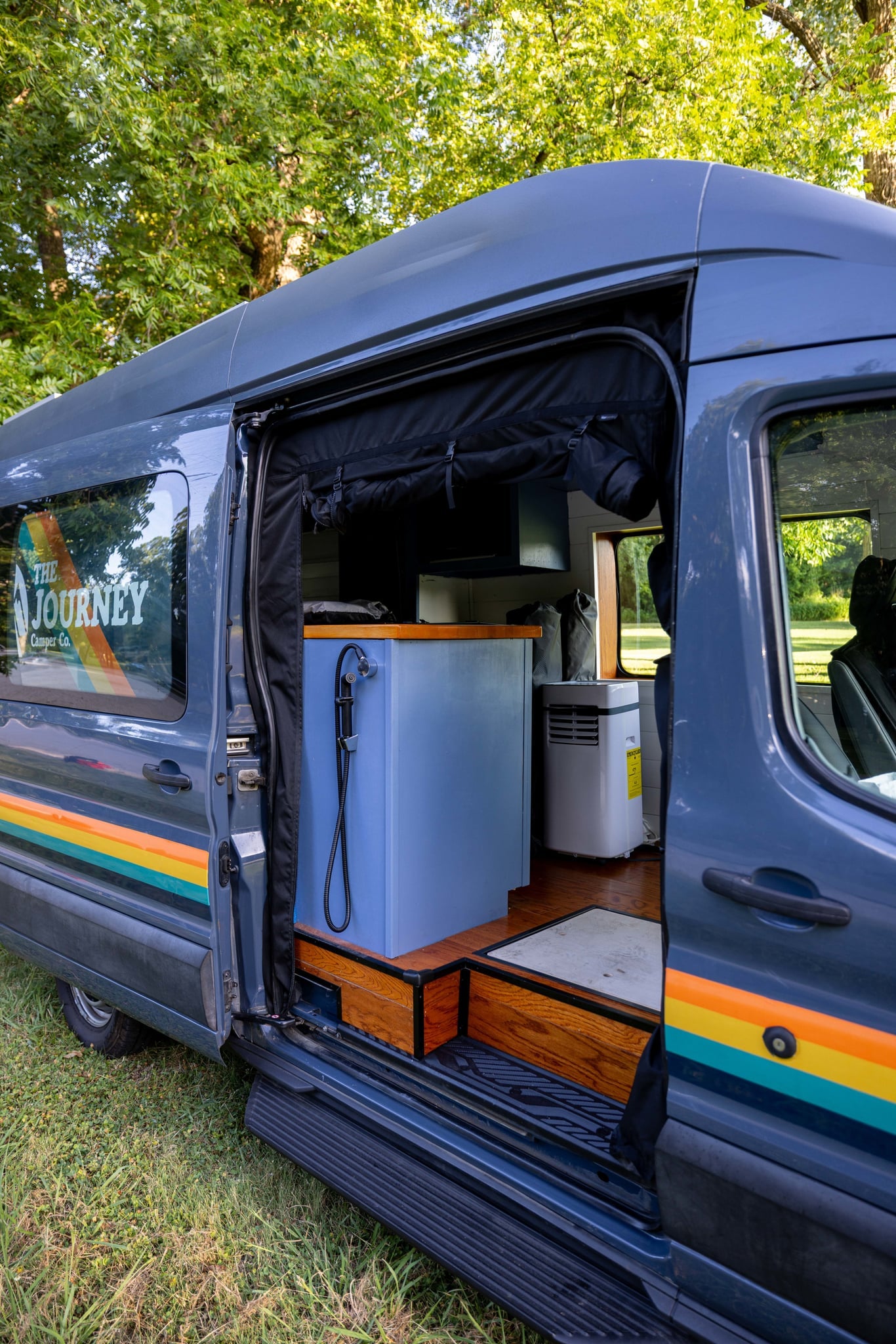  parked on grass among trees.|Rear of The Journey RV rental camper van with striped logo. Folding table