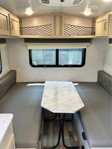 This RV rental shows a bright dinette with gray booth seating, a central table, overhead cabinets, and storage nets.