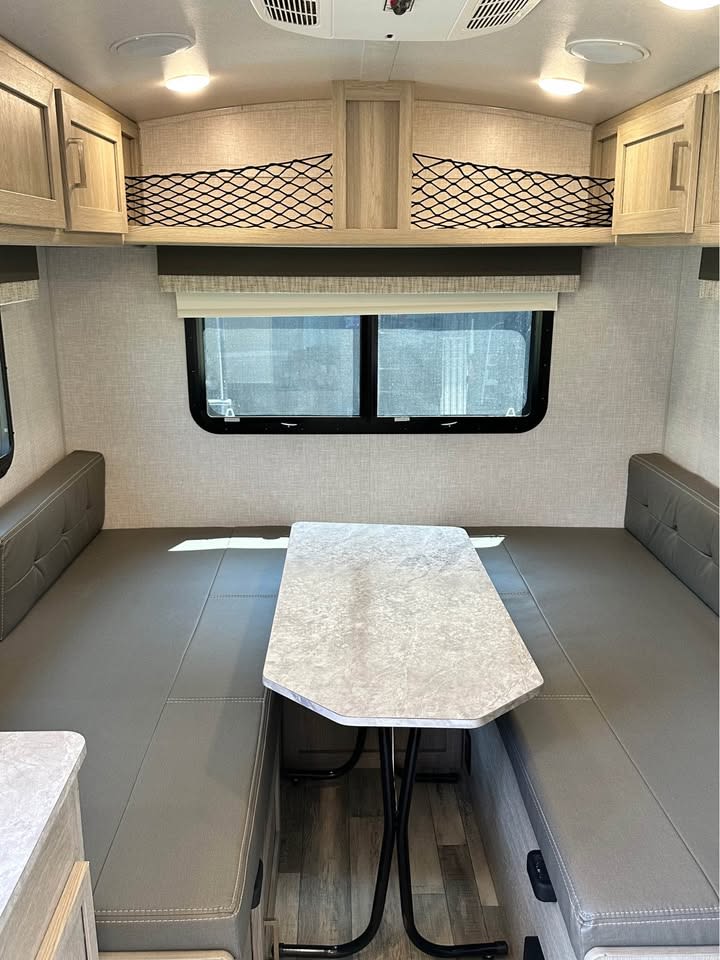 This RV rental shows a bright dinette with gray booth seating, a central table, overhead cabinets, and storage nets. — photo 8