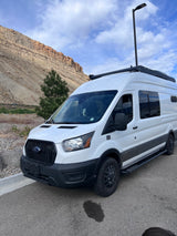A white RV rental van with a happy dog, parked on asphalt by a towering rocky mountain and blue sky. Ready for adventure!