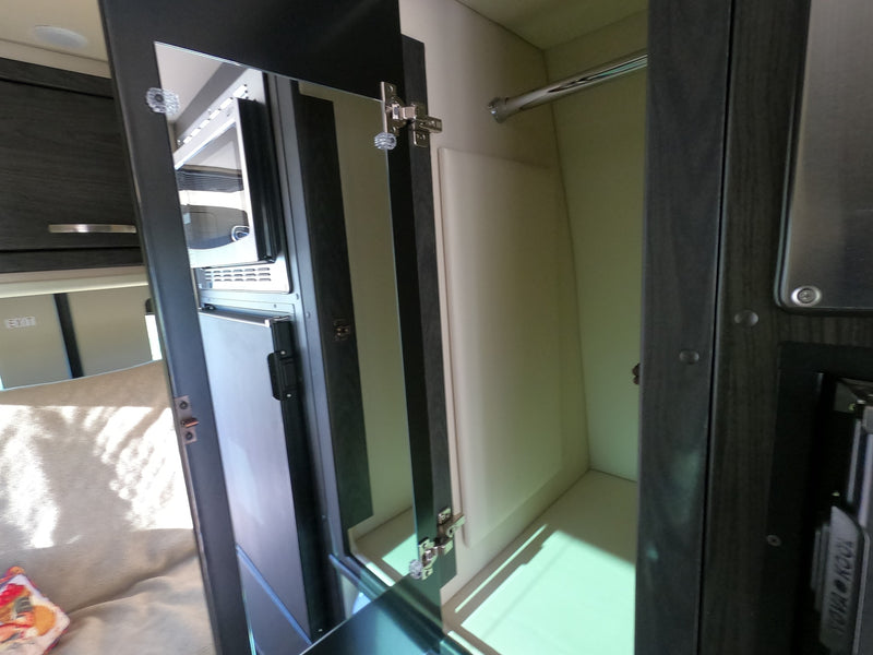 Compact rv rental interior with an open wardrobe and hanging bar, reflecting a view of the microwave, fridge, and seating area. — photo 11