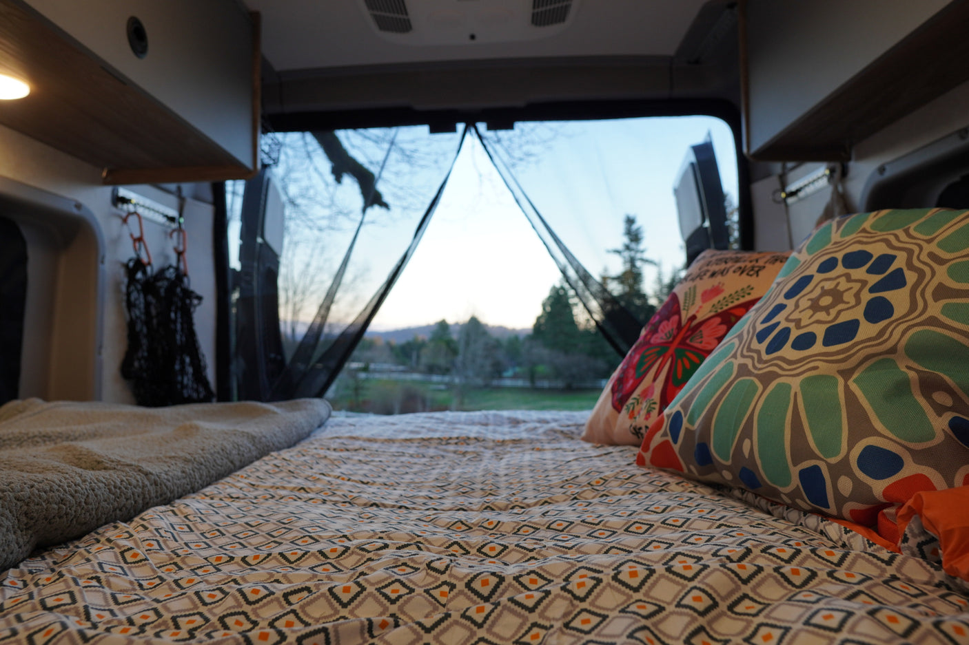 Cozy rv rental bed with patterned sheets and pillows, offering a scenic outdoor view through the open rear doors.