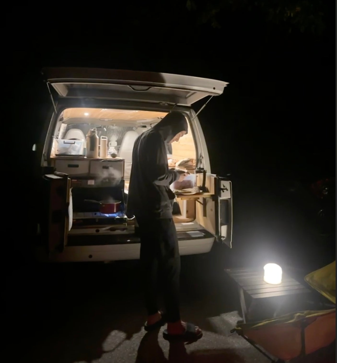 Nighttime scene: A person cooks from their RV rental, a camper van's back opens to an outdoor kitchen setup with a glowing lantern.