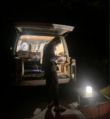 Nighttime scene: A person cooks from their RV rental, a camper van's back opens to an outdoor kitchen setup with a glowing lantern.