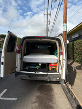A white van, possibly an rv rental, is seen with its back doors open, revealing a mattress for sleeping, parked by a "2 HR PARKING" sign.