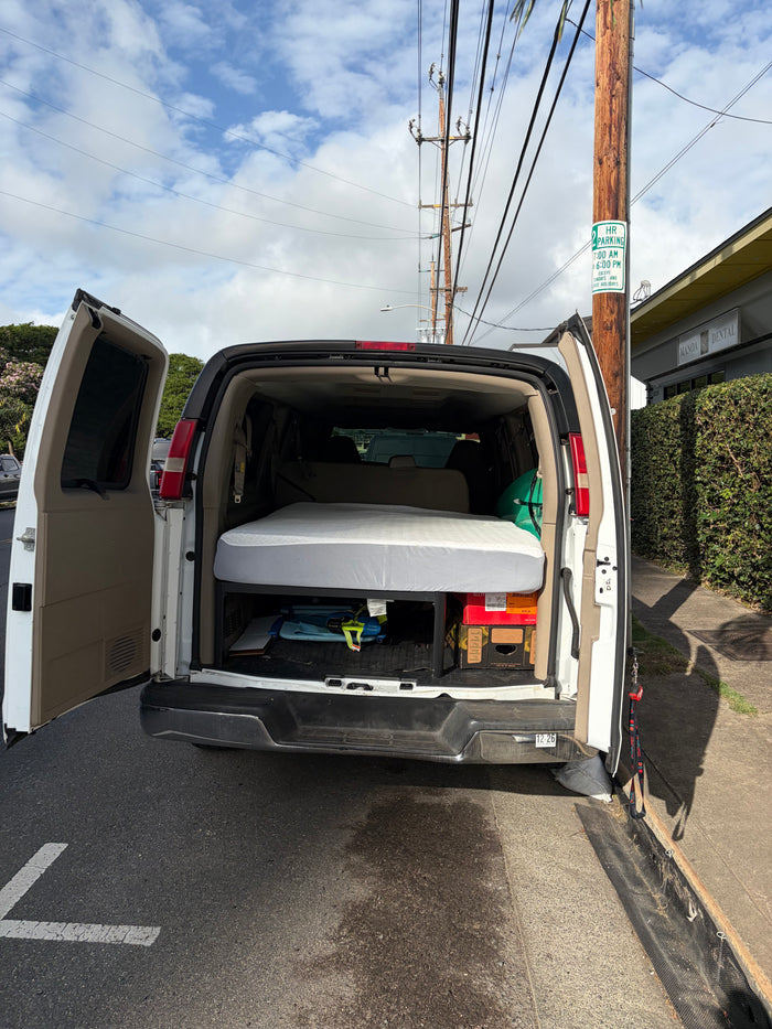 A white van, possibly an rv rental, is seen with its back doors open, revealing a mattress for sleeping, parked by a "2 HR PARKING" sign.