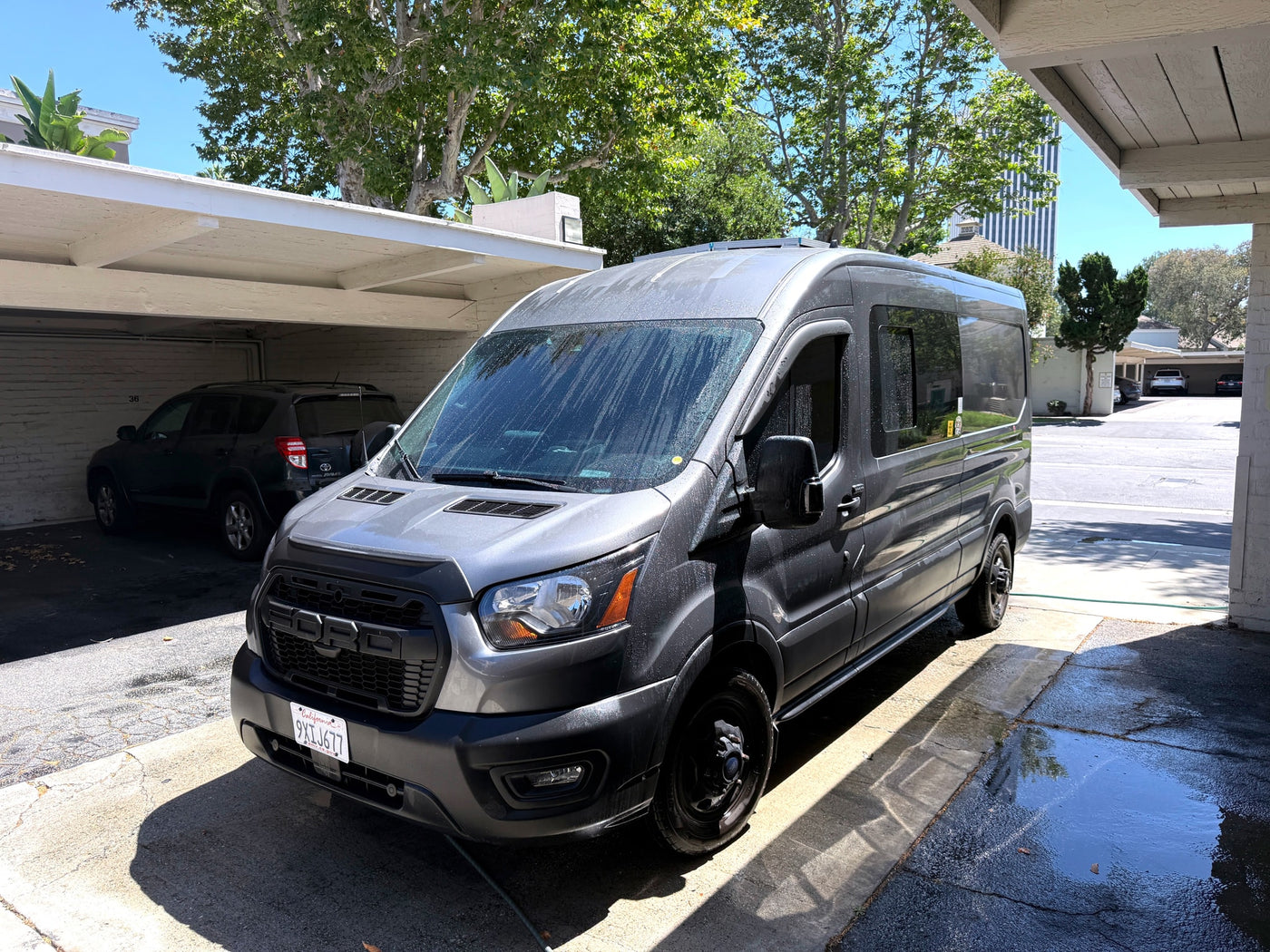 Gray Ford Transit camper van, an ideal rv rental, parked and wet in a sunlit carport area. — photo 2