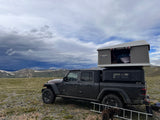 Dark grey Jeep Gladiator with a rooftop tent, an RV rental, overlooking snowy mountains under dramatic cloudy skies.