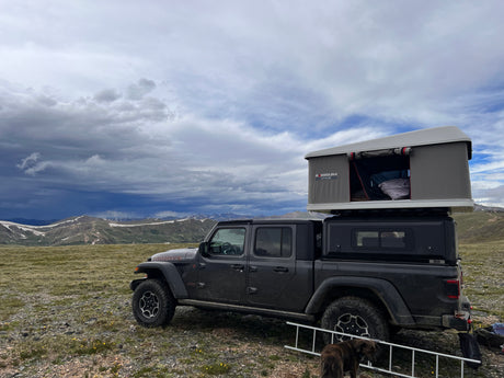 Dark grey Jeep Gladiator with a rooftop tent, an RV rental, overlooking snowy mountains under dramatic cloudy skies.