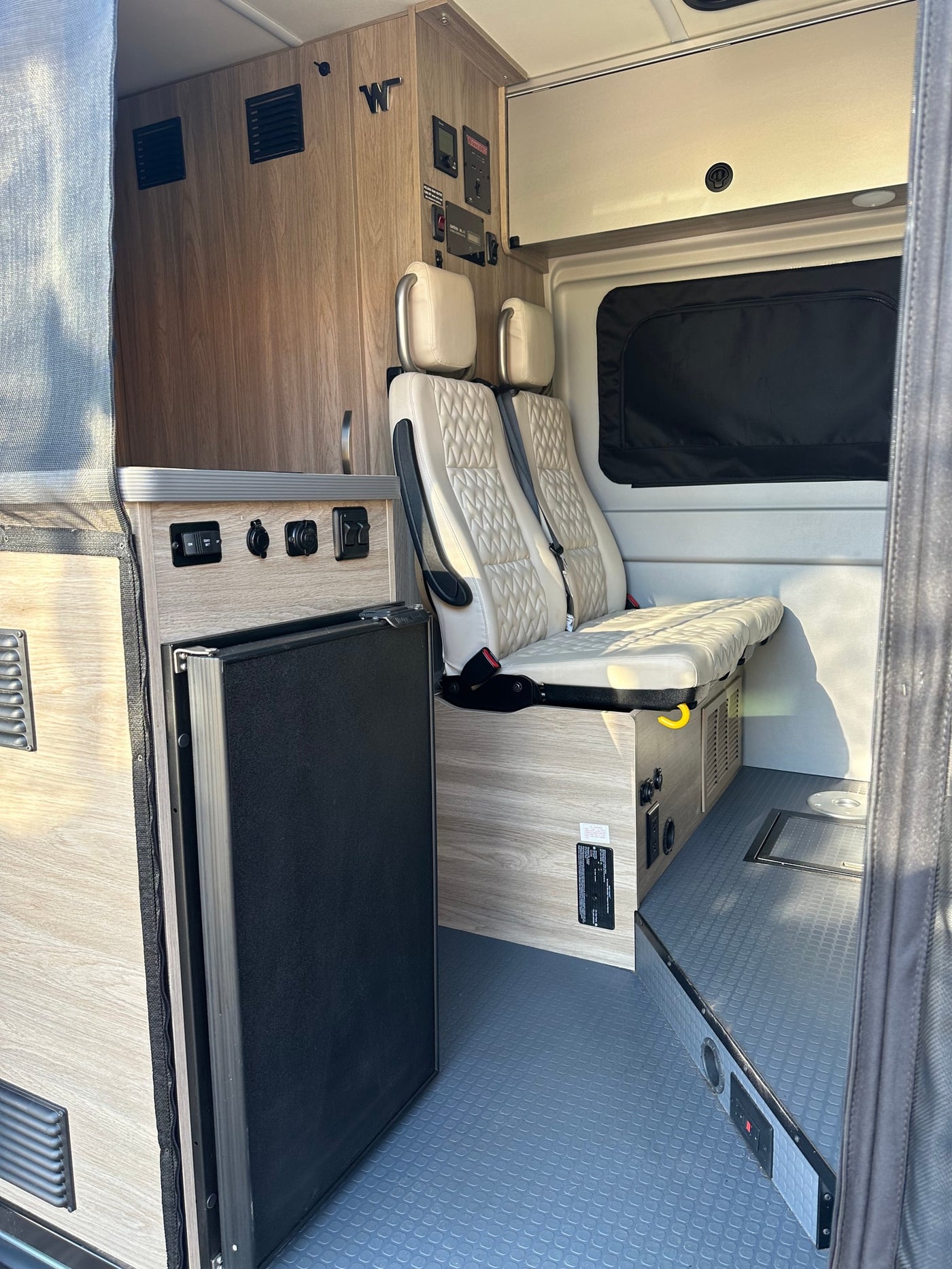 Inside this RV rental, light wood cabinetry, quilted passenger seats, a compact fridge, and control panel for adventure. — photo 13
