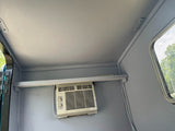 View of an RV rental's grey interior with a mounted AC unit, shelf, and window.