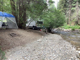 An RV rental van and screened tent by a river, nestled in a peaceful forest campsite.