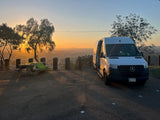 Golden hour illuminates an rv rental parked at a scenic overlook, offering stunning city views alongside a picnic table for travelers.