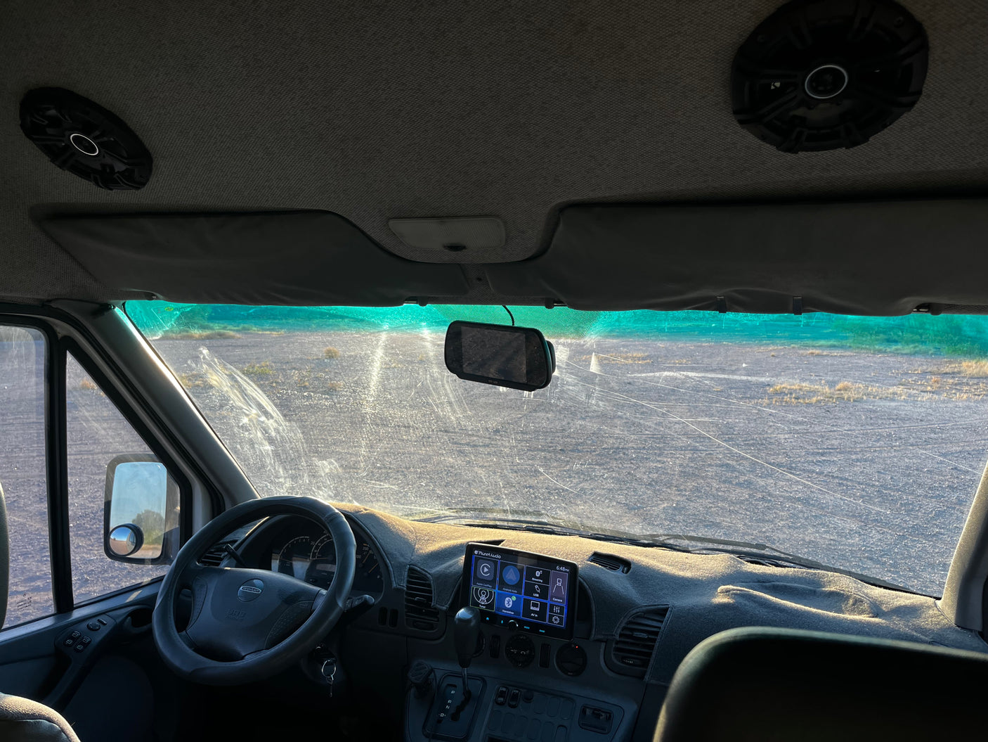 RV rental interior: Driver's view of the dashboard, steering wheel, and a dirty windshield looking out to an unpaved road.