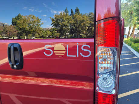 Close-up of a red Solis RV rental van, showing its unique sun logo, rear door handle, and clear taillight.